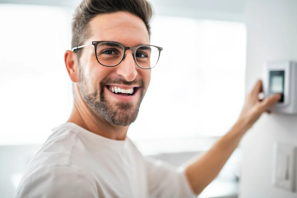 A smiling man demonstrating energy efficiency in HVAC by holding a thermostat in his home.