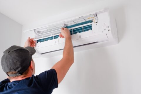 A technician in a baseball cap addresses common HVAC issues by servicing an indoor air conditioning unit, adjusting the internal components.