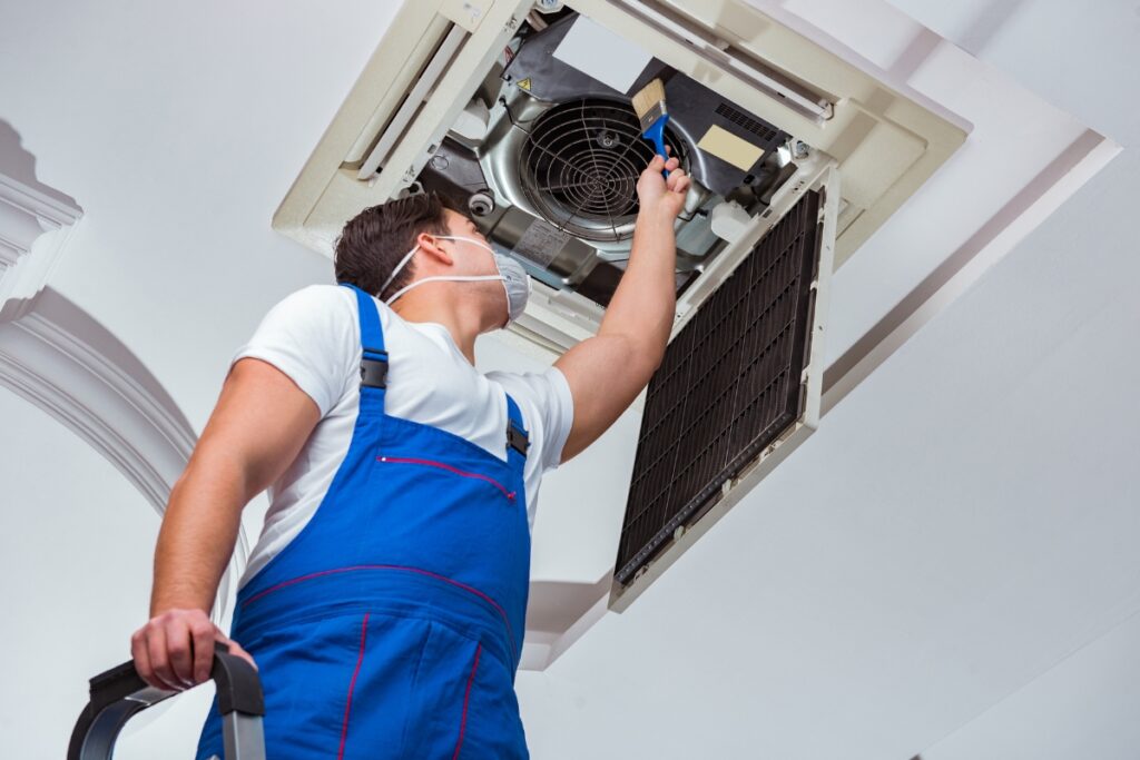 A technician in overalls and safety goggles addressing common HVAC issues while repairing an air conditioning unit on a ceiling.