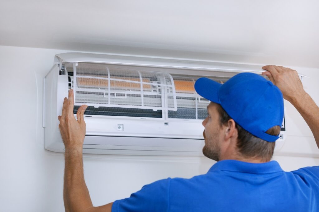 A technician in a blue uniform addressing common HVAC issues by servicing an indoor air conditioning unit and checking the filter.