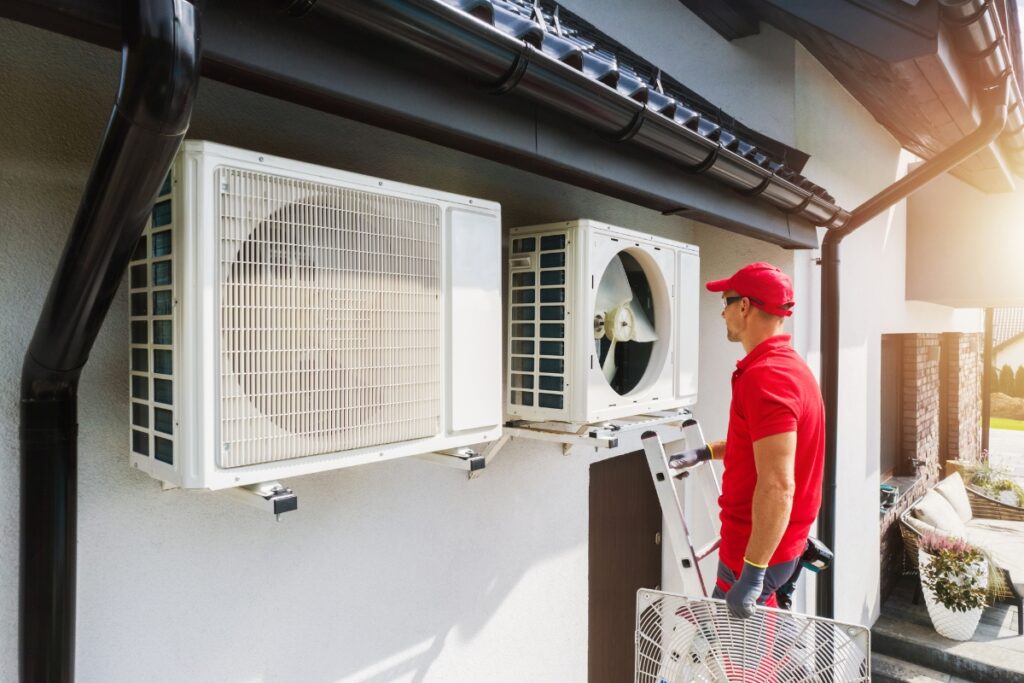 A technician in a red cap examines outdoor air conditioning units mounted on the side of a house for common HVAC issues.