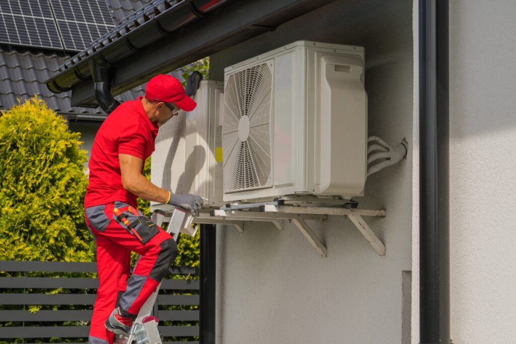 A technician in red overalls addressing common HVAC issues in an outdoor air conditioning unit attached to a house wall.