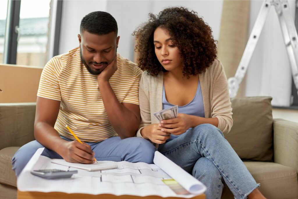 A couple reviewing architectural plans together while sitting on a couch, with the woman holding a small model house and discussing hvac upgrade costs.