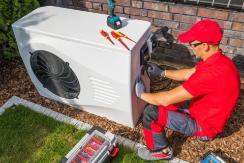 A technician in a red shirt and cap choosing the right HVAC system while servicing an outdoor air conditioning unit with tools nearby.