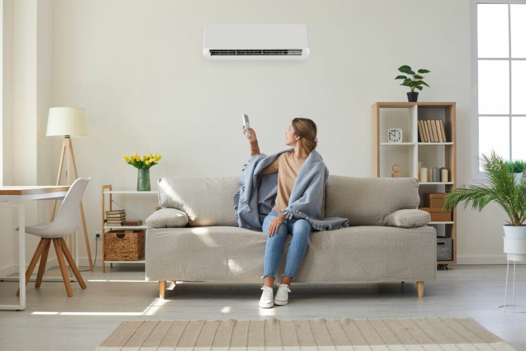 Woman sitting on a sofa in a bright living room, using a remote to adjust the air conditioner above her while choosing the right HVAC system.