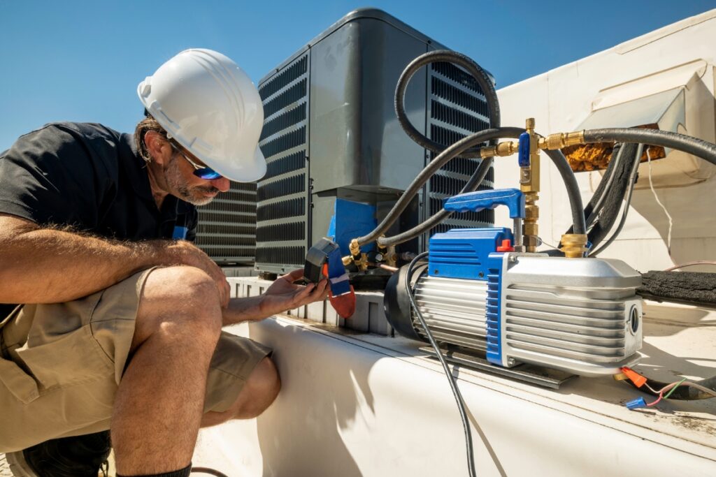 A technician in a white hard hat and black shirt works on an HVAC system on a roof under a clear blue sky. With various tools and equipment around him, he meticulously ensures everything adheres to HVAC building codes.