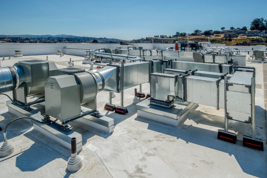 Rooftop view of commercial HVAC units with various ducts and vents against a backdrop of hills and a clear sky, all meticulously designed to meet HVAC building codes.