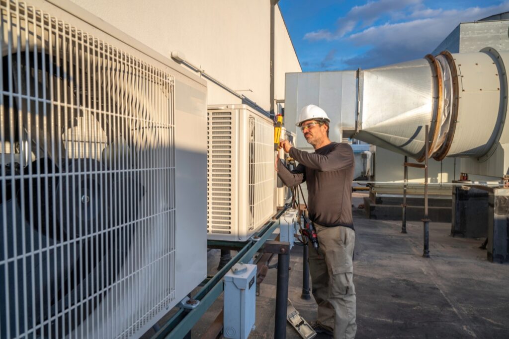 A worker in a hard hat and safety gear adheres to HVAC building codes while repairing an HVAC unit on a rooftop.