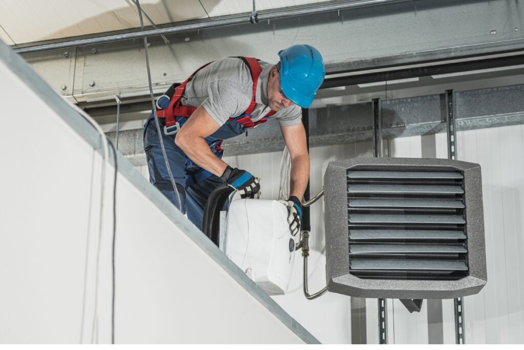 A worker in a blue helmet and safety gear is servicing a large ceiling-mounted ventilation unit, ensuring compliance with HVAC building codes.