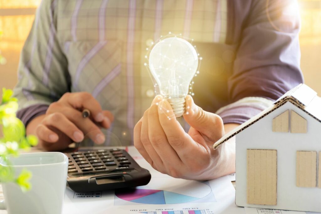 A person holding a glowing light bulb, seated at a desk with a calculator, documents, and a small model house, ponders how to make renewable energy at home.