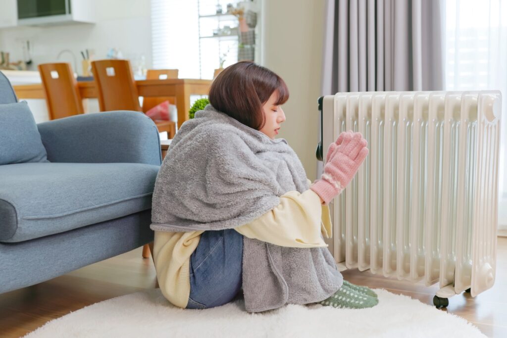 A person wrapped in a blanket and wearing gloves sits on the floor next to a heater in a cozy living room, enjoying the warmth and appreciating their home insulation's energy savings.