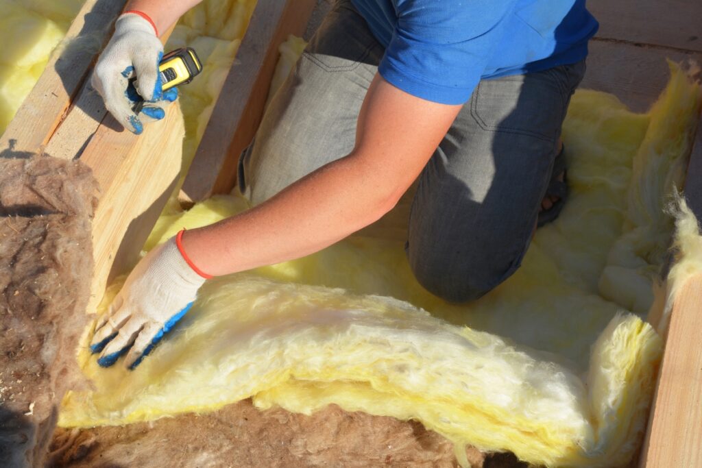 A person wearing gloves measures and installs yellow insulation material between wooden beams, ensuring optimal home insulation for energy savings.