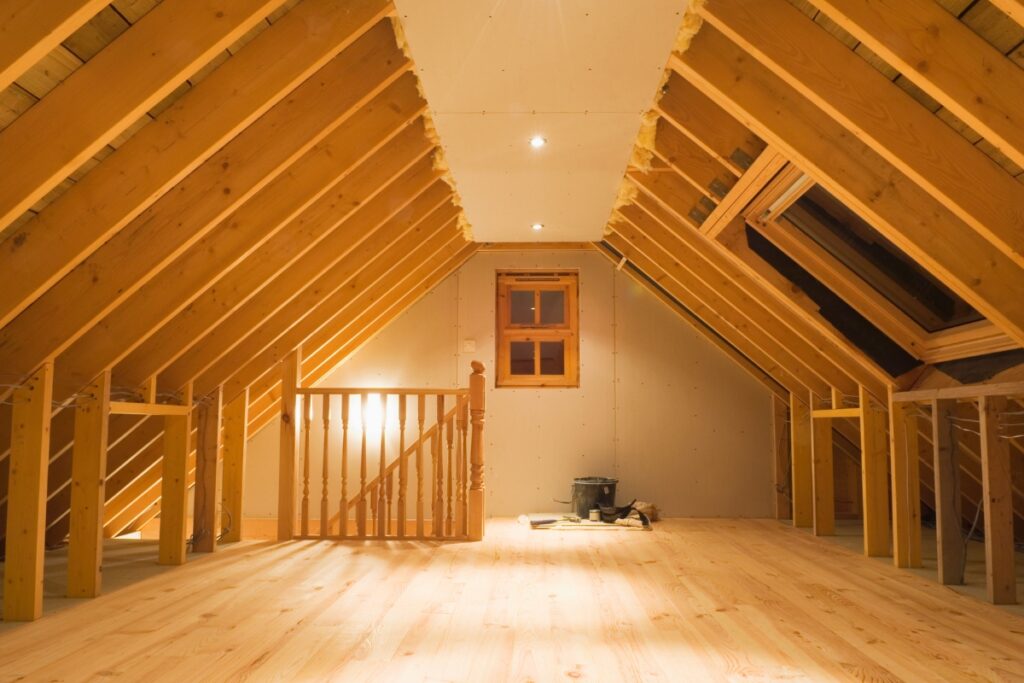 An unfinished attic with wooden beams, plywood floor, a staircase railing, and a small window at the far end. There are construction materials and tools scattered on the floor, ideal for installing home insulation to boost energy savings.