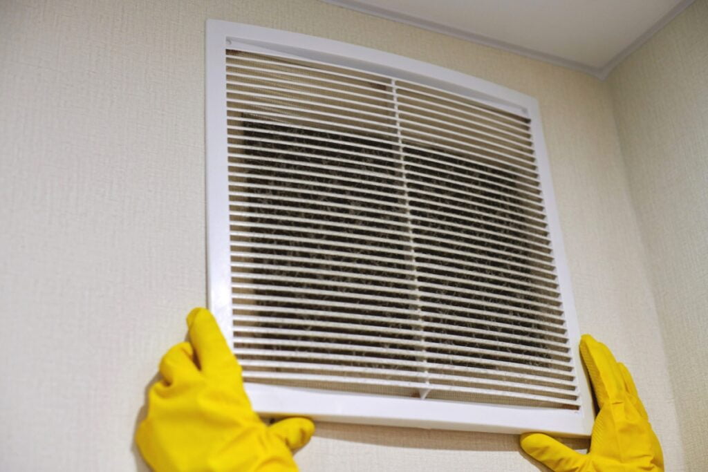 A person wearing yellow gloves removes a dirty air vent cover from a wall, contributing to improved home insulation and energy savings.