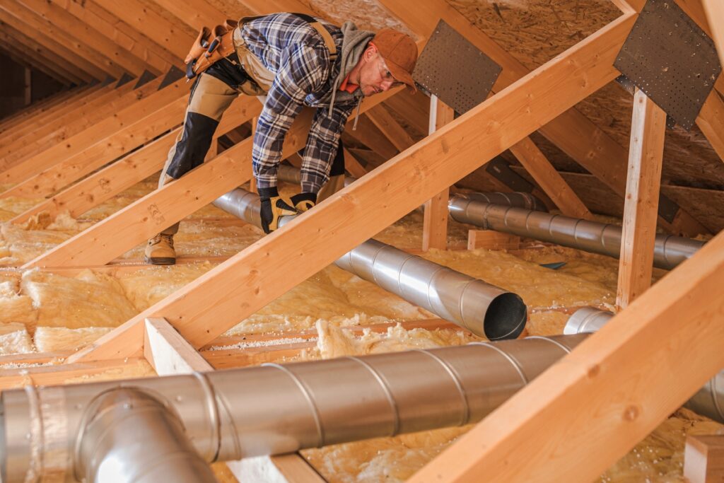 A worker in a beanie and flannel shirt installs home insulation in an attic, surrounded by wooden beams and metal ducts, ensuring future energy savings.