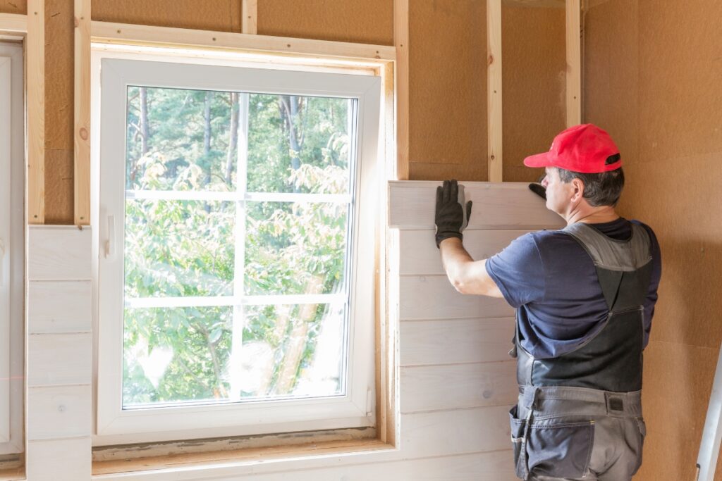 A person wearing a red cap and overalls is installing wooden paneling inside a partially constructed wall next to a window, ensuring better home insulation and energy savings.