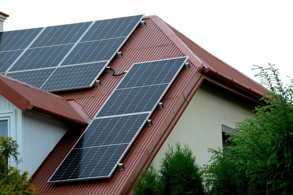 Solar panels, a key step in how to make renewable energy at home, are installed on the slanted red roof of a house, surrounded by green shrubs.