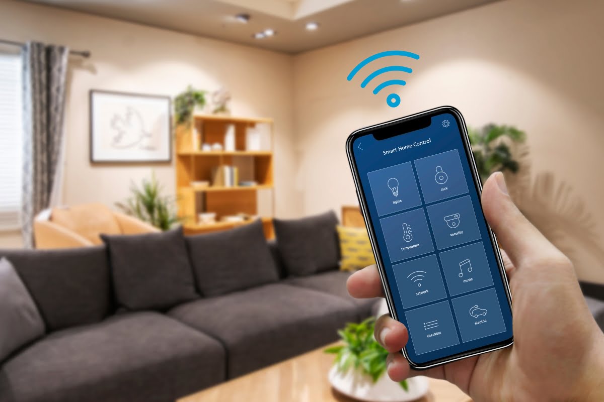 A hand holding a smartphone with a home automation app displayed, controlling various smart home HVAC functions. The background shows a modern living room with a couch, bookshelf, and indoor plants.