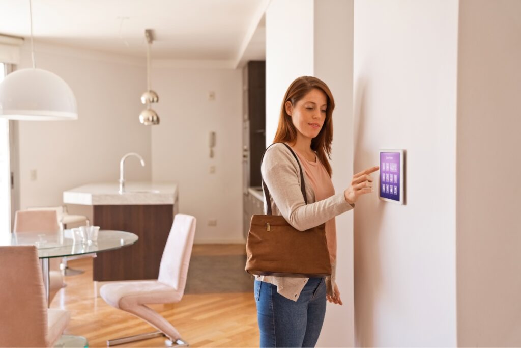 Woman using a touchscreen panel on the wall of a modern kitchen and dining area, adjusting the smart home HVAC system, while wearing a brown bag over her shoulder.