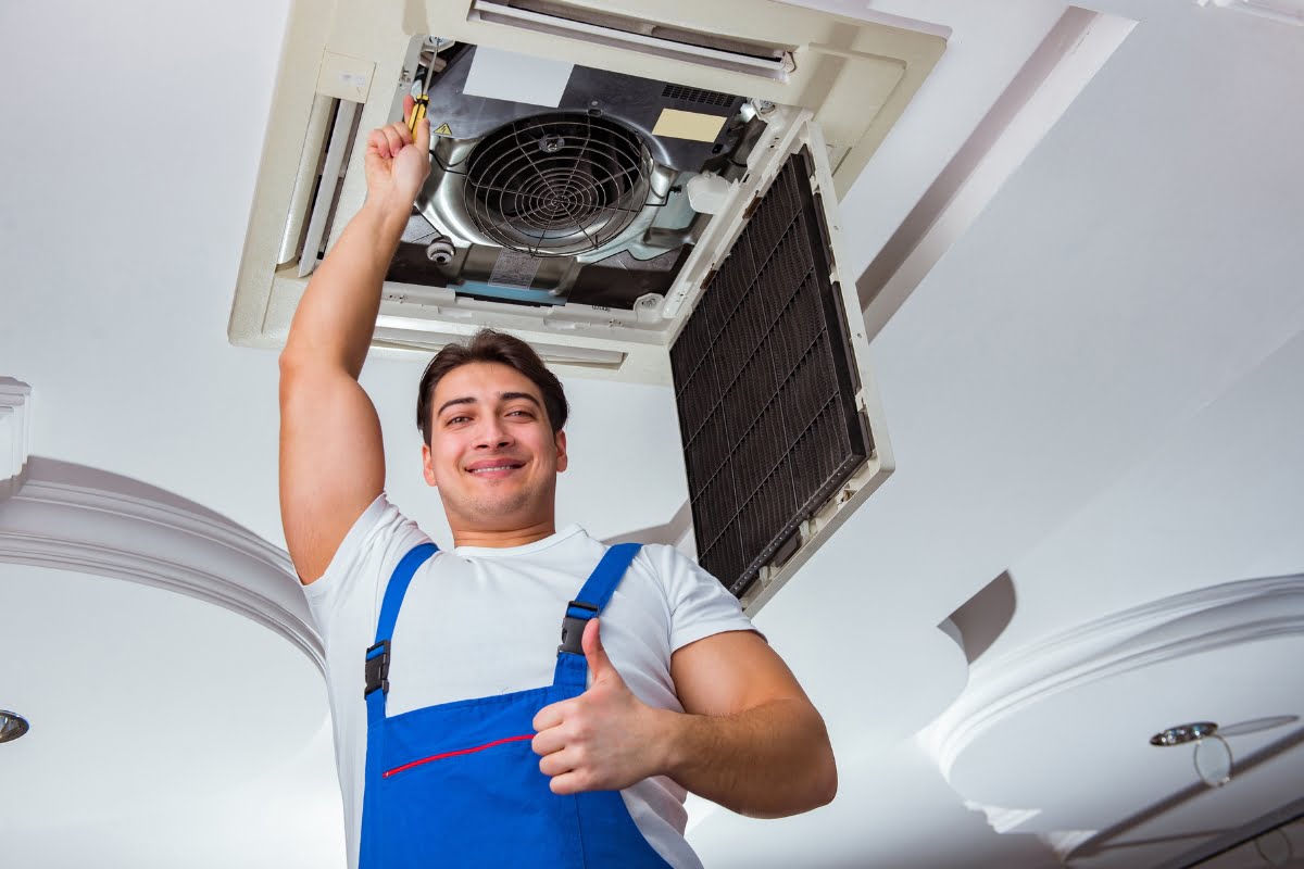 A technician in blue overalls services a ceiling-mounted HVAC unit, giving a thumbs-up to ensure a healthy home environment.