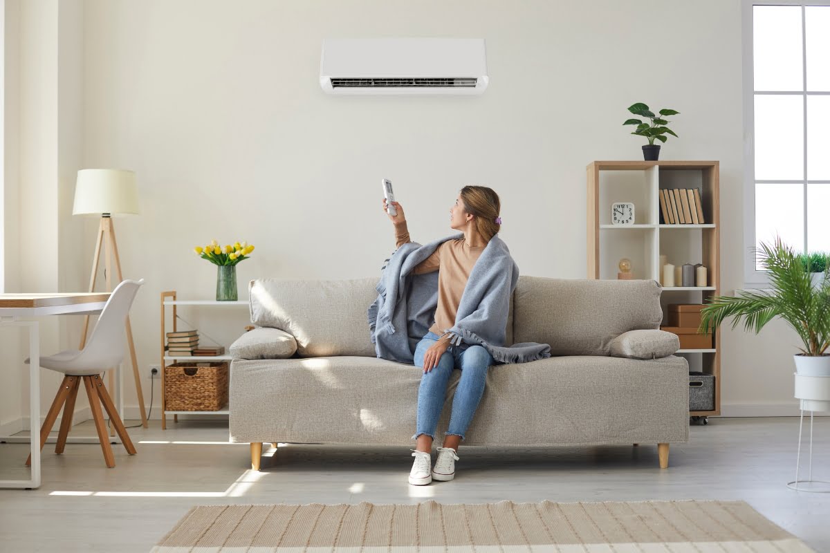 A woman sits on a sofa covered with a blanket, pointing a remote control at a wall-mounted air conditioner in her bright, modern living room, ensuring she enjoys the benefits of an efficient HVAC system for a healthy home environment.