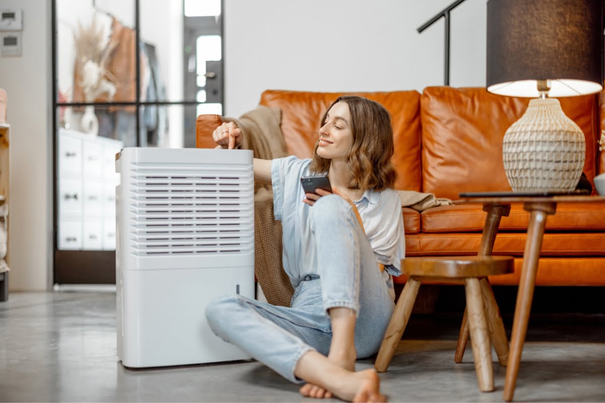 A woman sitting on the floor in a living room adjusts a control on a white air purifier, ensuring her healthy home environment. An orange sofa and a lamp add warmth to the background.