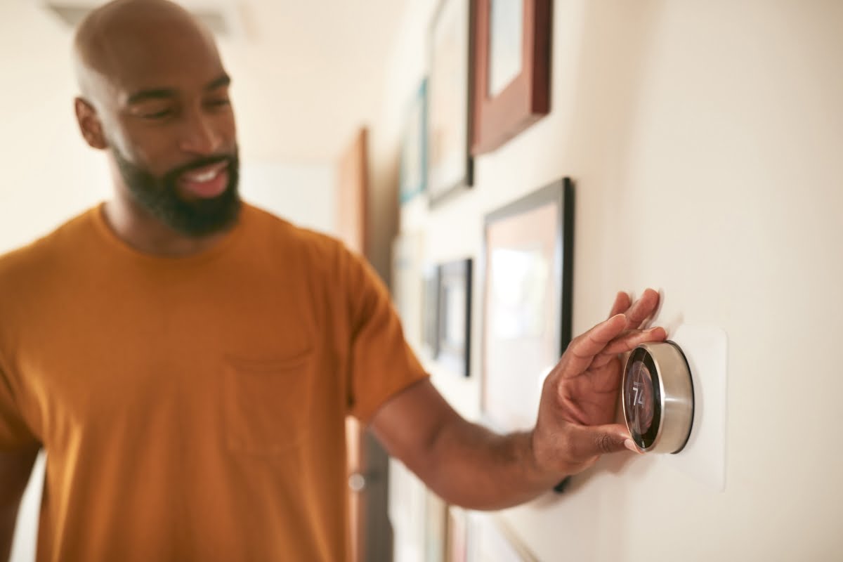 A man in a mustard-colored shirt adjusts a modern thermostat on the wall, reflecting his commitment to efficient HVAC control in an era of climate change.