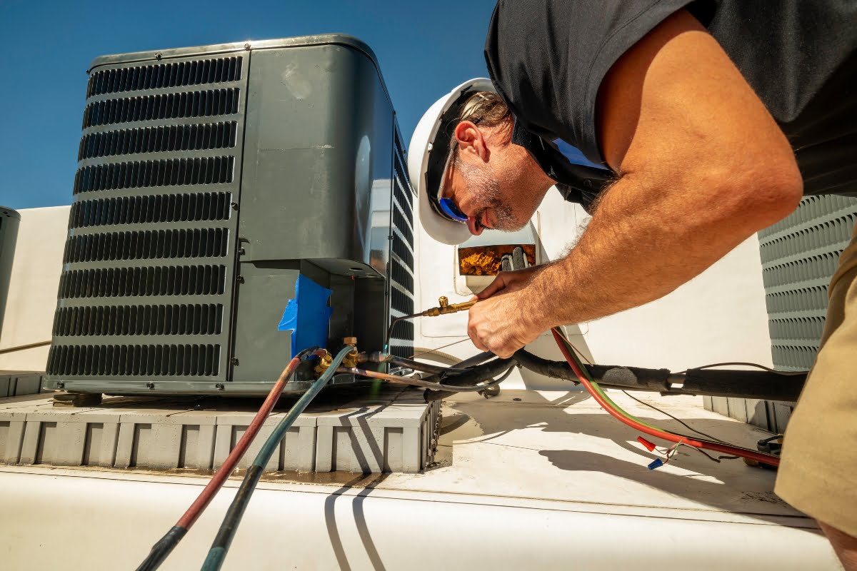 A technician wearing a hard hat and sunglasses works on an HVAC unit on a rooftop, adjusting various cables and components in bright sunlight, ensuring optimal performance to address climate change challenges.