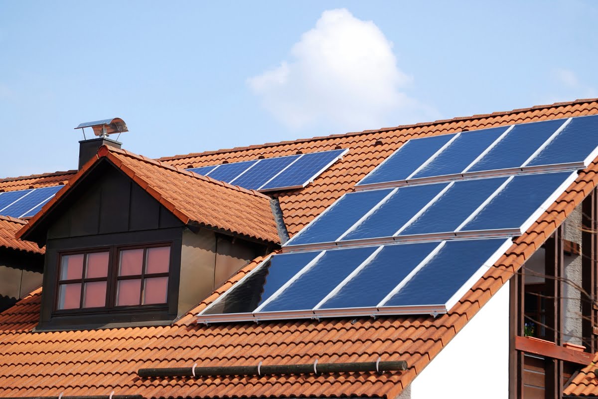 A house with terracotta roof tiles and multiple solar panels installed on the roof stands against a clear sky with a few clouds, reflecting its eco-friendly design in the face of climate change.