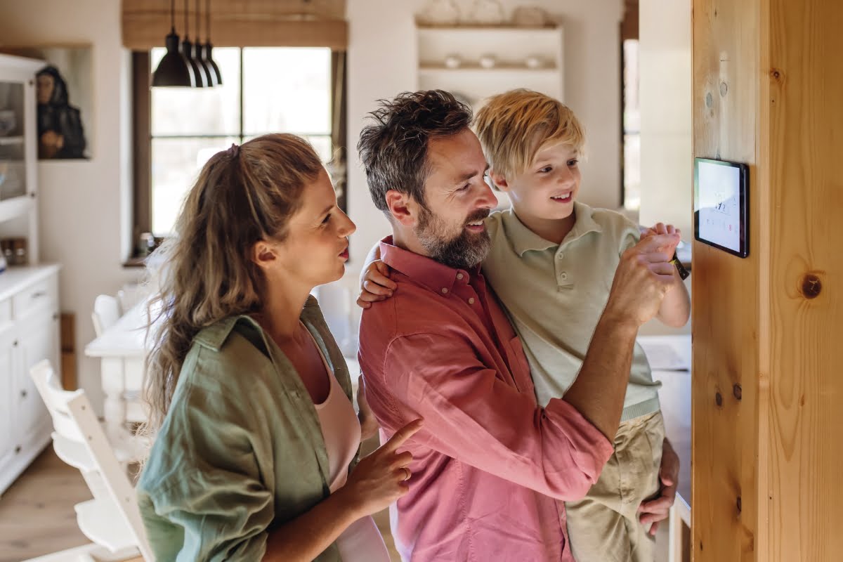 A woman, man, and child are standing in a cozy home, interacting with a digital screen mounted on a wooden wall. The man is holding the child, who is eagerly pointing at the screen displaying HVAC settings that contribute to climate change awareness.