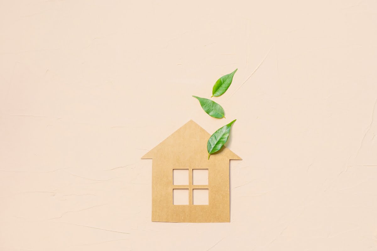 A cardboard cutout of a house with green leaves emerging from the chimney, subtly highlighting climate change, against a light beige background.