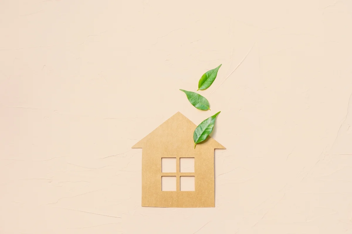 A cardboard cutout of a house with green leaves emerging from the chimney, subtly highlighting climate change, against a light beige background.