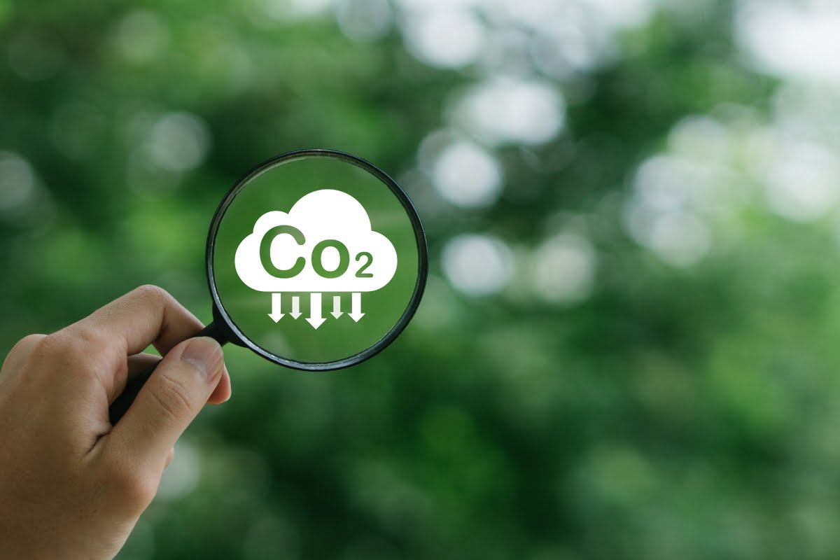 A hand holding a magnifying glass with a CO2 cloud icon, indicating carbon dioxide reduction, against a blurred green foliage background. This image underscores the role HVAC systems play in addressing climate change.