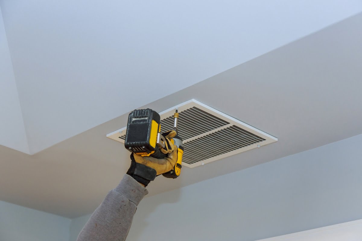 A person wearing gloves uses a yellow drill to install or remove a vent cover on a ceiling during an HVAC inspection.