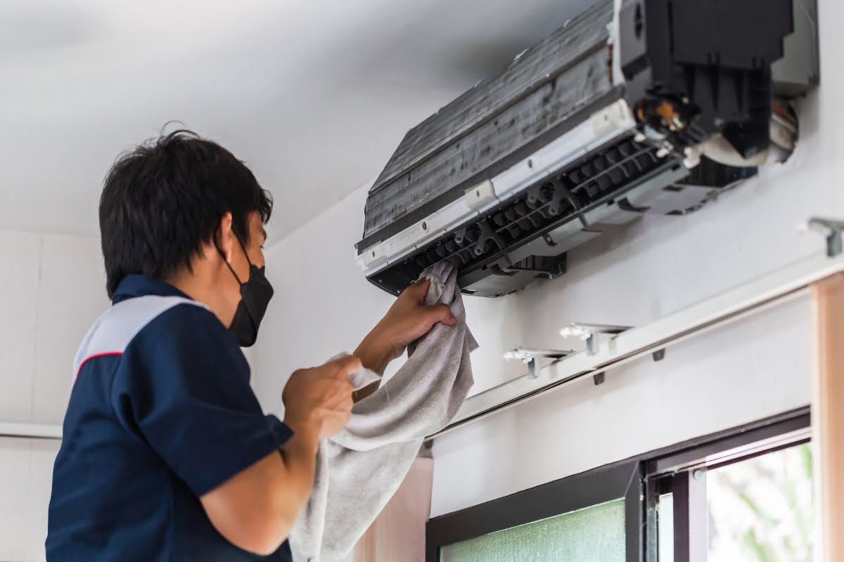 A person wearing a mask meticulously cleans the interior of a wall-mounted air conditioning unit with a cloth, ensuring it meets HVAC standards for real estate inspection.