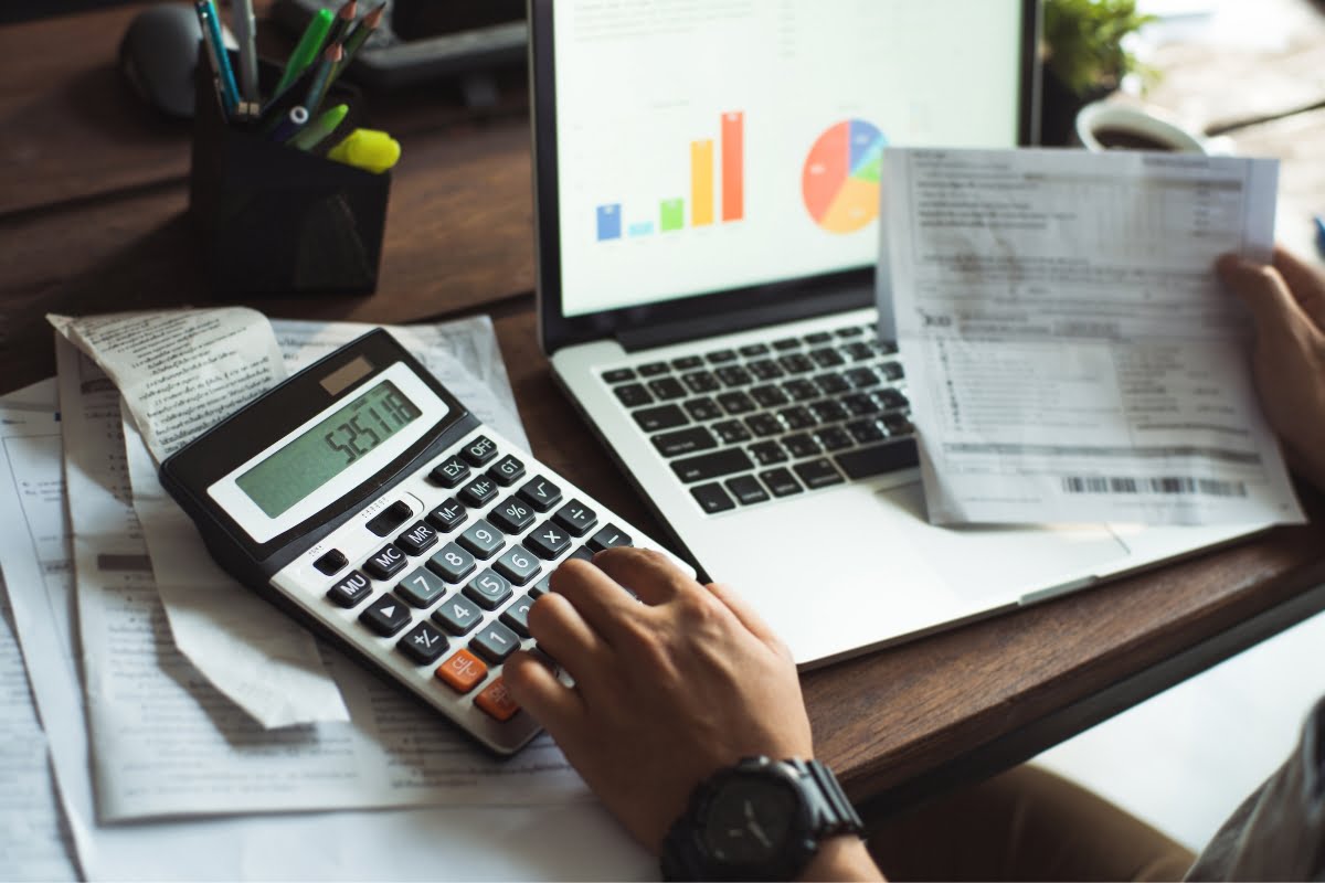 Individual working at a desk with a laptop, calculator, and various financial documents, analyzing data charts and figures related to HVAC real estate inspection.