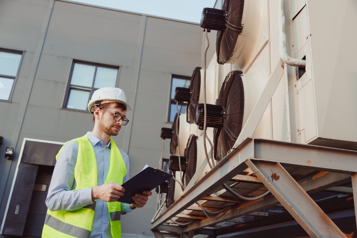 A worker wearing a white hard hat and a yellow safety vest holds a clipboard while conducting an HVAC inspection on large units mounted on the side of a building, ensuring everything meets real estate standards.