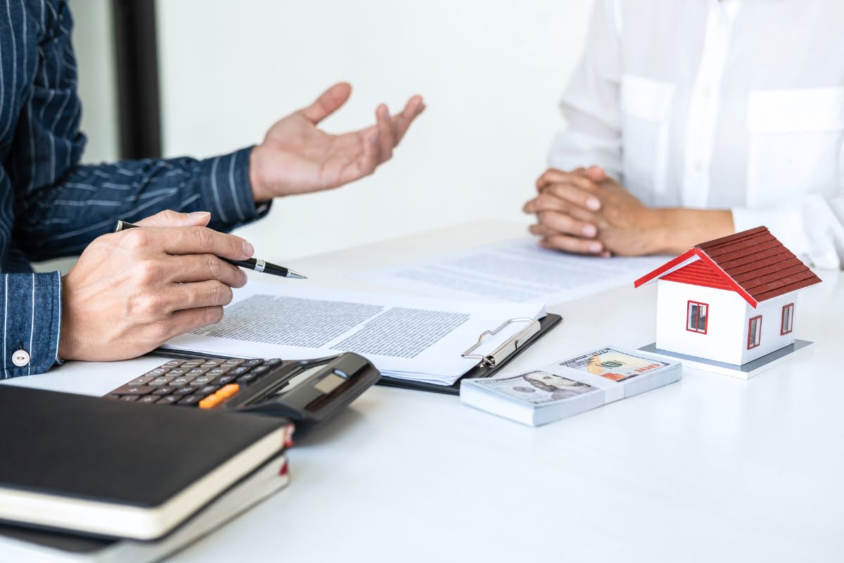 Two people seated at a table, discussing documents. The table has a calculator, a notebook, a small house model, and a stack of money—likely reviewing details from a recent real estate inspection.