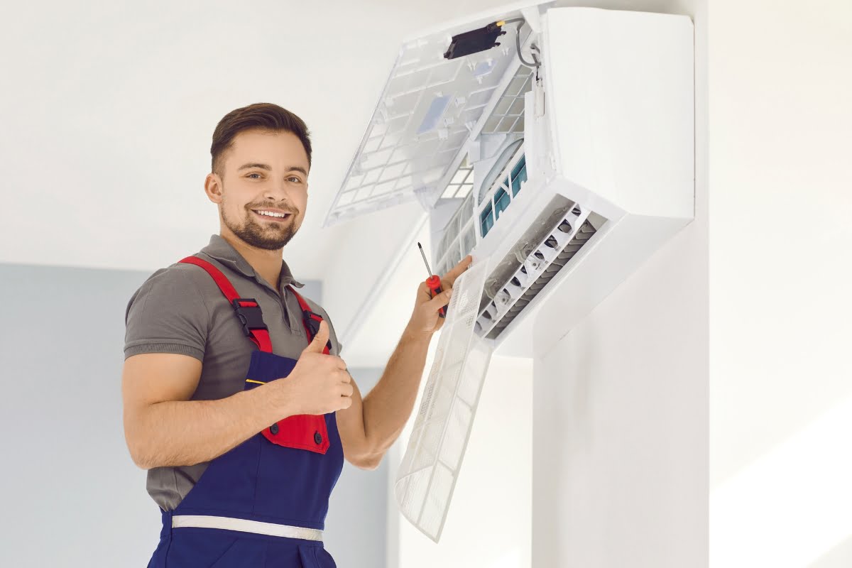 A technician wearing a grey shirt and blue overalls gives a thumbs up while servicing an air conditioning unit mounted on a light-colored wall, during an HVAC real estate inspection.