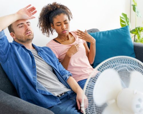 A couple enjoying the air conditioning while sitting on a couch with a fan.