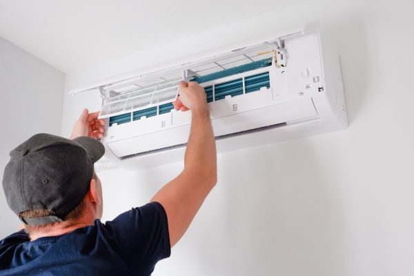 A technician in a baseball cap addresses common HVAC issues by servicing an indoor air conditioning unit, adjusting the internal components.