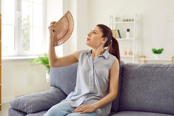 A woman sits on a gray couch in a well-insulated living room, using a handheld fan to cool herself, enjoying the energy savings.