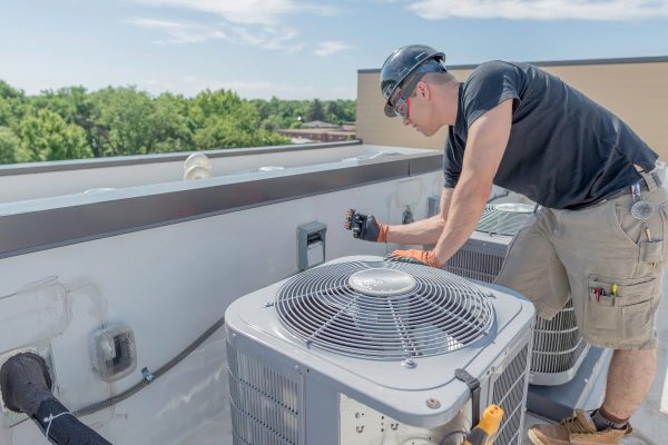 A technician wearing a hard hat and gloves is inspecting an air conditioning unit on a rooftop on a clear day, ensuring it complies with HVAC building codes.